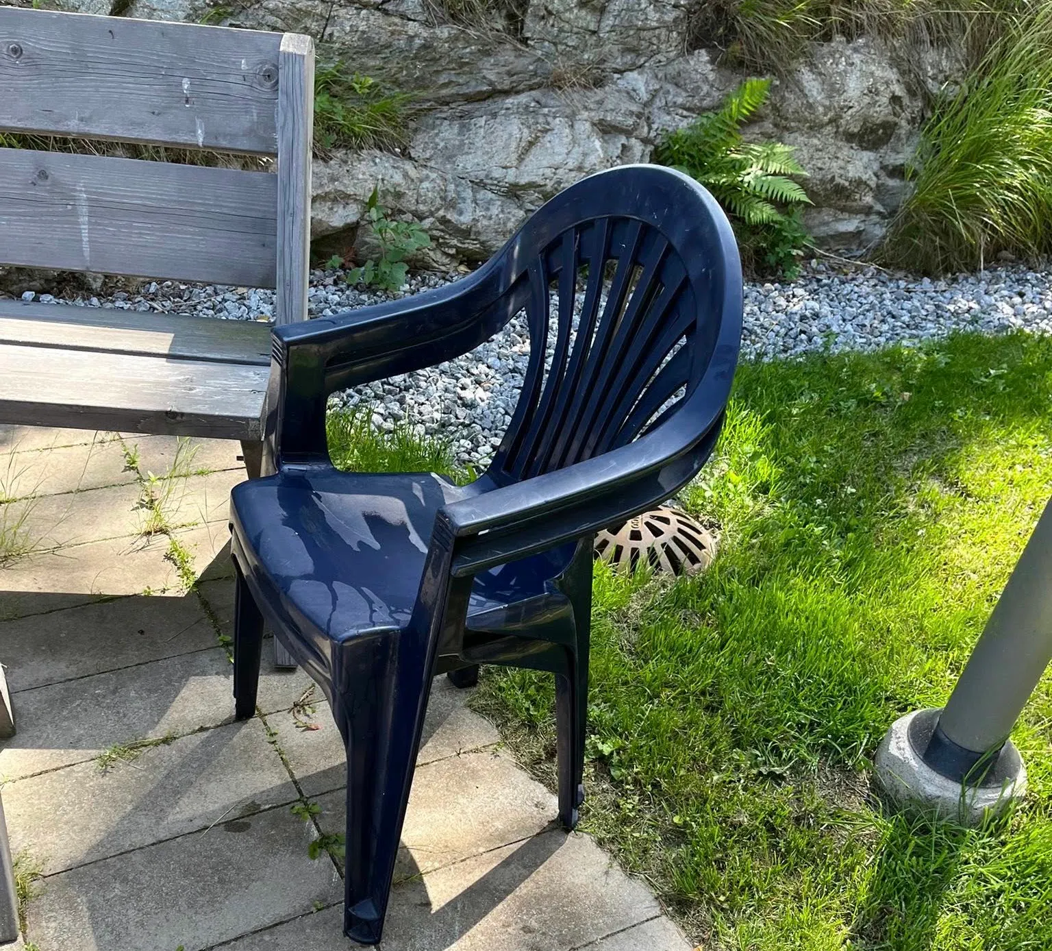 Två staplade blå plaststolar på stenplattor mellan en träbänk och en gräsmatta.